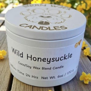 May include: A white candle tin with a lid featuring a dog face design and the words "SWAN WICK CANDLES". The label reads "Wild Honeysuckle" and "Coco/Soy Wax Blend Candle". The net weight is 6oz.
