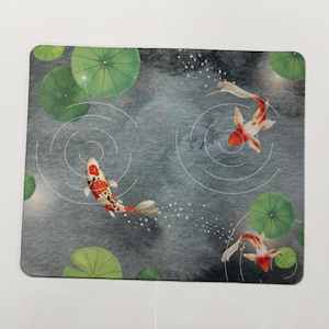 May include: A rectangular mat with a watercolour design of a pond with three koi carp. The fish are orange and white, swimming among green lily pads. Concentric circles suggest ripples in the water.