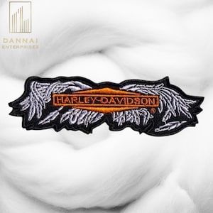 May include: Black embroidered patch with the words "HARLEY-DAVIDSON" in orange, framed by a black border and white wing-like designs. The patch is rectangular and features the brand's logo.