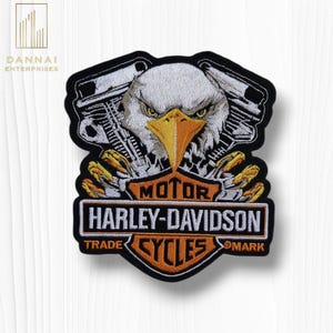 May include: Embroidered patch featuring a detailed eagle head with a yellow beak and piercing eyes, set against a backdrop of motorcycle engine components. The patch includes the text "MOTOR HARLEY-DAVIDSON CYCLES".