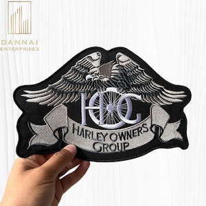 May include: Black embroidered patch featuring two eagles above the letters "HOG" and a motorcycle wheel design. The text "HARLEY OWNERS GROUP" is displayed on a banner below. The patch is held against a white background.