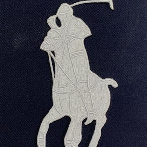 May include: A white embroidered patch featuring a polo player on a horse, set against a navy blue background. The rider is depicted in a classic pose, holding a mallet. The patch is likely for apparel or accessories.