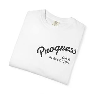 Puede incluir: Una camiseta blanca doblada con la frase negra «Progress Over Perfection». El texto está en escritura cursiva y letras de bloque. La camiseta tiene cuello redondo y es de material suave.
