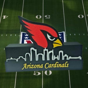 Könnte beinhalten: Ein schwarz-graues Arizona Cardinals-Logo mit einem roten Kardinalvogel und gelbem Schnabel. Das Logo zeigt eine Skyline-Silhouette und die gelben Worte "Arizona Cardinals". Der Hintergrund ist ein grünes Fußballfeld.