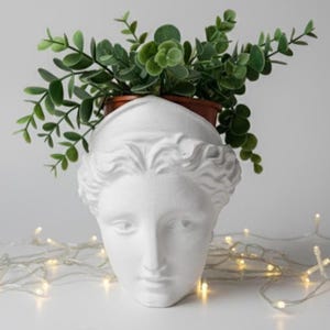 Könnte beinhalten: Ein weißer Pflanztopf in Form einer klassischen Büste mit einem ruhigen Gesicht, der einen kleinen braunen Topf mit grünen Blättern hält. Der Pflanztopf steht auf einer weißen Oberfläche, mit einer Lichterkette im Hintergrund.