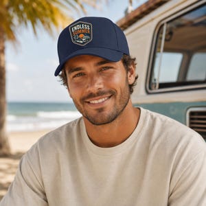 May include: A navy blue baseball cap featuring a shield-shaped patch on the front. The patch displays a sunset scene with a palm tree, a van, and the text ENDLESS SUMMER in white and orange lettering against a dark background.