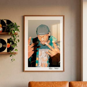 May include: A framed print featuring a person wearing a cap with the word "WOLFNESS" and a graphic tee. The artwork is displayed above a brown leather sofa. Vinyl records and a plant are on a shelf.