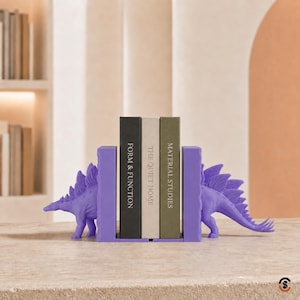 May include: A pair of purple stegosaurus dinosaur bookends supporting three books titled Form & Function, The Quiet Home, and Material Studies. The bookends feature a solid purple color and a detailed dinosaur shape with plates along the back.