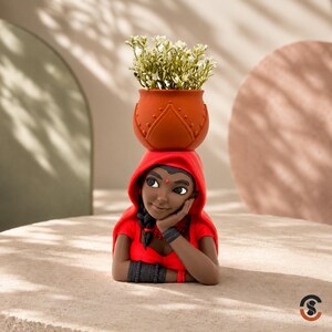 May include: A decorative figurine of a person with dark skin wearing a bright red hooded garment and dark bracelets, supporting a terracotta-colored pot on their head. The pot contains a small arrangement of white flowers with green stems.