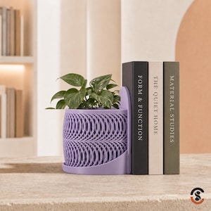 May include: A lavender-colored planter with a repeating circular lattice pattern, integrated into a bookend design. The planter holds a green leafy plant and is positioned next to three books titled Form & Function, The Quiet Home, and Material Studies.