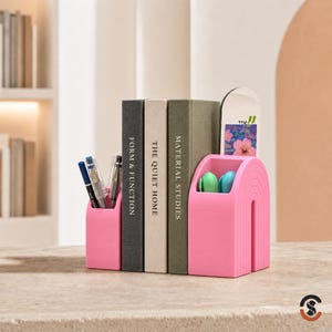 May include: A pink desk organizer with an arched design holding pens and markers, positioned alongside three books titled Form & Function, The Quiet Home, and Material Studies, with a small rectangular card featuring a floral graphic.
