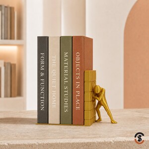 May include: A set of four books with spines labeled Form & Function, The Quiet Home, Material Studies, and Objects in Place, held in position by a gold-colored bookend featuring a stylized human figure pushing against a brick wall structure.