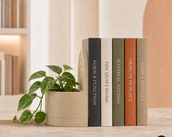 May include: A beige arched planter with a green pothos plant positioned next to five books titled Form & Function, The Quiet Home, Material Studies, Objects in Place, and Simple Rituals, featuring spines in black, white, olive, terracotta, and tan.