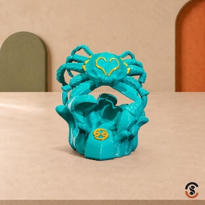 May include: A turquoise crab figurine featuring a gold heart-shaped emblem on its back and a gold Cancer zodiac symbol on the front base. The crab is depicted with raised claws and textured legs, resting on a sculpted base with a smooth, matte finish.