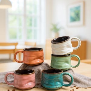 Peut inclure: Ensemble de tasses en céramique de différentes couleurs : rose, orange, turquoise, vert et blanc. Chaque tasse présente une surface texturée et un intérieur marron foncé. Les tasses sont disposées sur une table en bois, avec des grains de café éparpillés.