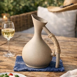 Peut inclure: Une cruche en céramique beige avec une poignée enroulée de corde est posée sur un tissu bleu. Un verre de vin blanc et une assiette de salade sont sur une table en bois. Guirlande lumineuse et chapeau de paille en arrière-plan.