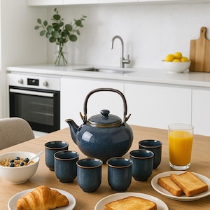 May include: A blue ceramic tea set with a teapot and six cups, each with a gold rim, arranged on a wooden table. A croissant, toast, and a bowl of cereal are also on the table, along with a glass of orange juice.