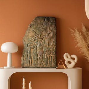 May include: A weathered, rectangular relief sculpture with hieroglyphs and figures, possibly Egyptian. The artwork is in shades of green and brown, depicting a scene with a sun and figures. The sculpture is displayed on a white table with other decorative items.