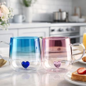 Peut inclure: Deux tasses en verre transparent avec des bases en forme de cœur. L'une est bleue, l'autre rose. Les deux ont des poignées transparentes. Les tasses sont sur une surface blanche avec des articles de petit-déjeuner et un vase de fleurs en arrière-plan.
