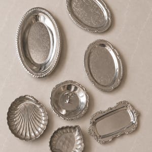 May include: A collection of seven silver-toned metal dishes and trays in various shapes, including oval, rectangular, shell-shaped, and leaf-shaped designs. Each piece features decorative edges and polished surfaces with intricate embossed detailing.