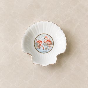May include: A white porcelain shell-shaped dish featuring a central circular illustration of two pink flamingos standing in water with reeds. The dish has a scalloped edge with a thin gold trim and the word Florida printed below the flamingos.
