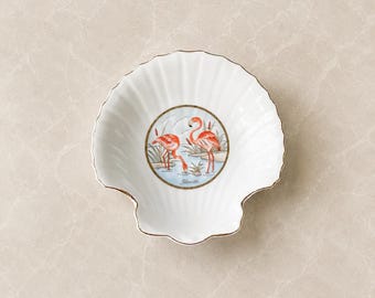 May include: A white porcelain shell-shaped dish featuring a central circular illustration of two pink flamingos standing in water with reeds. The dish has a scalloped edge with a thin gold trim and the word Florida printed below the flamingos.
