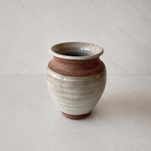 Peut inclure: Un petit vase en céramique avec un corps arrondi et un bord large et évasé. La pièce présente un design bicolore avec un vernis blanc cassé lisse et brillant sur la section centrale, tandis que le bord et la base révèlent l'argile brute.