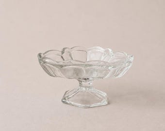 May include: A clear glass pedestal dish featuring a scalloped rim and a shallow, wide bowl. The base of the dish is hexagonal in shape, supporting a short, thick stem that connects to the bowl. The glass is transparent with a smooth, reflective finish.