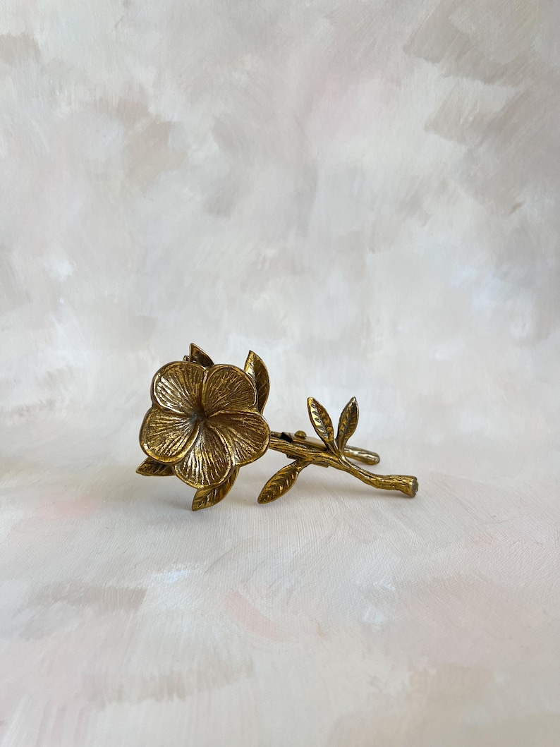 May include: A detailed, antique-style brass flower and stem. The flower has five petals and is surrounded by leaves. The stem extends to the right, with additional leaves. The piece is set against a soft, painted background.