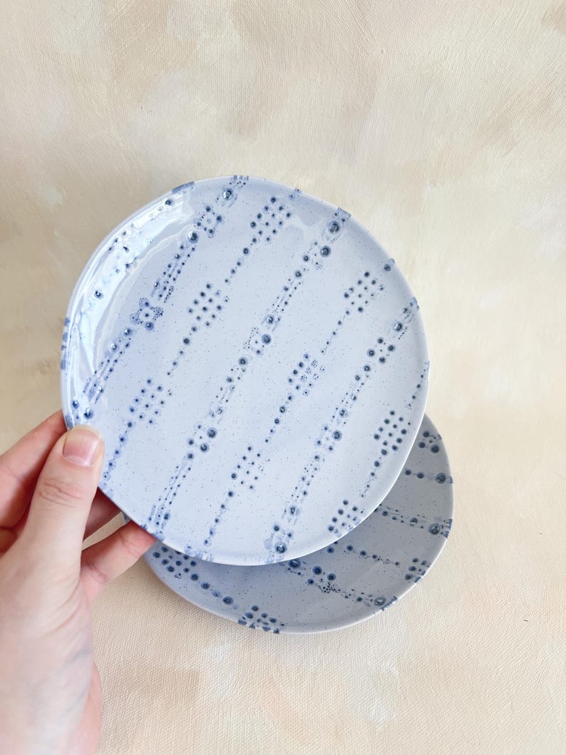 Puede incluir: Dos platos de cer&aacute;mica azul claro con un patr&oacute;n de puntos en azul m&aacute;s oscuro. Los platos son redondos y tienen una textura moteada. Un plato se sostiene ligeramente por encima del otro, mostrando el dise&ntilde;o.