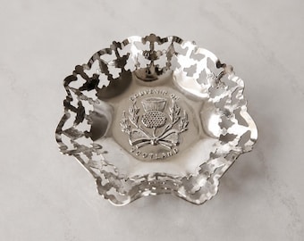 May include: A silver-toned metal dish featuring a decorative scalloped, pierced rim. The center of the dish displays an embossed thistle emblem with a bow and the text "SOUVENIR OF SCOTLAND" arched around the design in a circular arrangement.