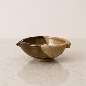 May include: A ceramic bowl with a pour spout and a small handle. The exterior and interior feature a two-tone glaze, with one side in a dark brown finish and the other side in a speckled beige finish. The bowl has a circular base and a wide rim.