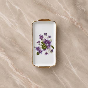 Peut inclure: Un plateau rectangulaire en porcelaine blanche orné d'une illustration florale centrale de violettes pourpres avec des feuilles vertes. Le plateau présente une finition lisse et brillante, bordée d'un fin liseré doré métallique sur les bords.