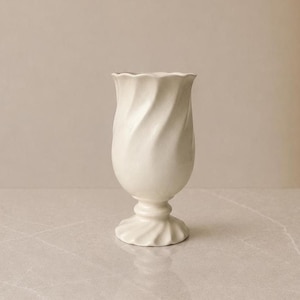 Peut inclure: Un vase en céramique de couleur crème présentant un motif en spirale ou torsadé distinct sur tout son corps. Le vase possède un large bord festonné et repose sur une base circulaire en forme de piédestal qui rappelle le motif torsadé.