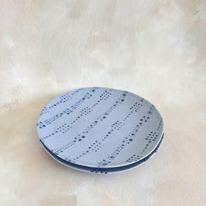 Puede incluir: Dos platos de cer&aacute;mica azul claro apilados. Cada plato presenta un patr&oacute;n de peque&ntilde;os puntos y guiones azul oscuro dispuestos en l&iacute;neas diagonales. El fondo es de color beige claro.