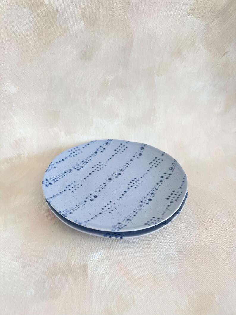 Puede incluir: Dos platos de cer&aacute;mica azul claro apilados. Cada plato presenta un patr&oacute;n de peque&ntilde;os puntos y guiones azul oscuro dispuestos en l&iacute;neas diagonales. El fondo es de color beige claro.