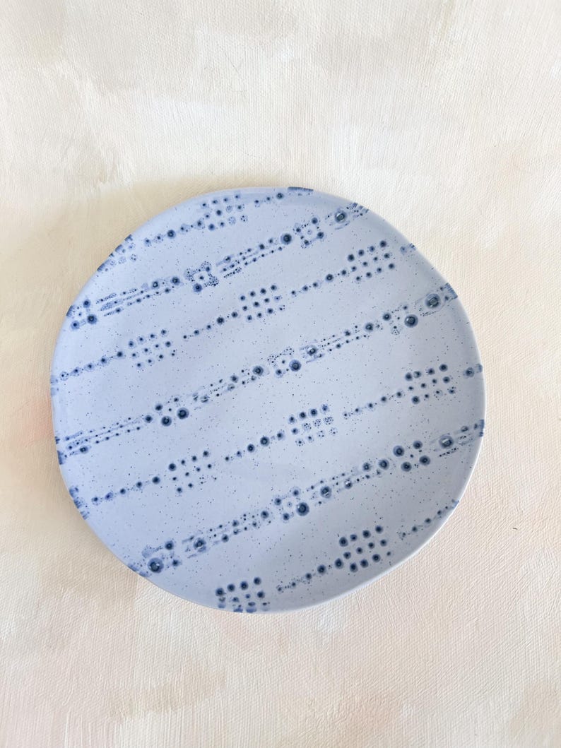 Puede incluir: Un plato redondo de cer&aacute;mica azul claro con un patr&oacute;n de puntos azul oscuro. El dise&ntilde;o presenta filas de peque&ntilde;os puntos y puntos m&aacute;s grandes agrupados, creando una apariencia texturizada. El plato tiene un borde ligeramente irregular.