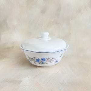 May include: A white ceramic bowl with a lid. The bowl has a blue floral pattern and a scalloped rim with a blue accent line. The lid has a small, rounded handle. A classic piece for serving or storage.