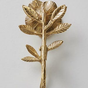 May include: A detailed, gold-colored metal floral decorative object. The design features a flower with multiple petals, leaves, and a stem. The object is likely a decorative element for home decor or a similar application.