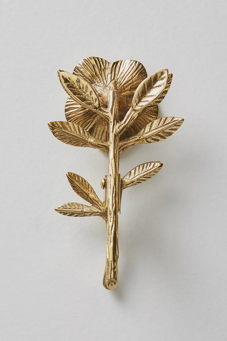 May include: A detailed, gold-colored metal floral decorative object. The design features a flower with multiple petals, leaves, and a stem. The object is likely a decorative element for home decor or a similar application.
