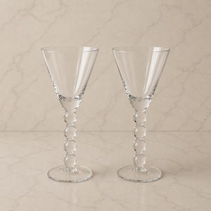 May include: A pair of clear glass goblets featuring a wide, tapered bowl and a stem composed of four stacked spherical glass beads. Each glass stands on a circular base, providing a stable foundation for the elegant, transparent drinking vessel.