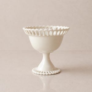 May include: A white ceramic footed bowl with a decorative, openwork rim and base. The bowl has a smooth, glossy finish and a classic, elegant design. The bowl is suitable for serving desserts or displaying small items.