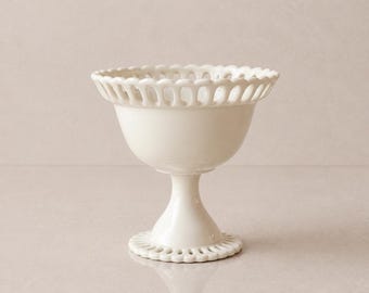 May include: A white ceramic footed bowl with a decorative, openwork rim and base. The bowl has a smooth, glossy finish and a classic, elegant design. The bowl is suitable for serving desserts or displaying small items.