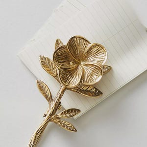 May include: A gold-colored metal flower-shaped object with a stem and leaves, resting on a lined notepad. The flower has detailed petals and a textured surface, suggesting a decorative or artistic piece.