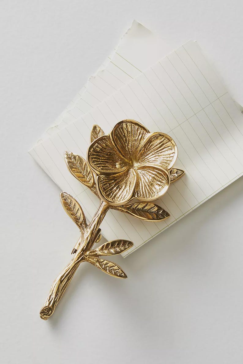 May include: A gold-colored metal flower-shaped object with a stem and leaves, resting on a lined notepad. The flower has detailed petals and a textured surface, suggesting a decorative or artistic piece.