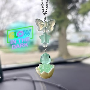 May include: A car charm featuring a butterfly, star, and turtle design. The charm is suspended from a silver chain. The turtle is inside a yellow and white egg. The text "GLOW IN THE DARK" is visible in the background.