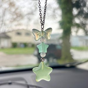 May include: A car charm featuring a chain with a butterfly, star, and bead design. The charm has a light green and clear color scheme. The charm is hanging in front of a car dashboard.