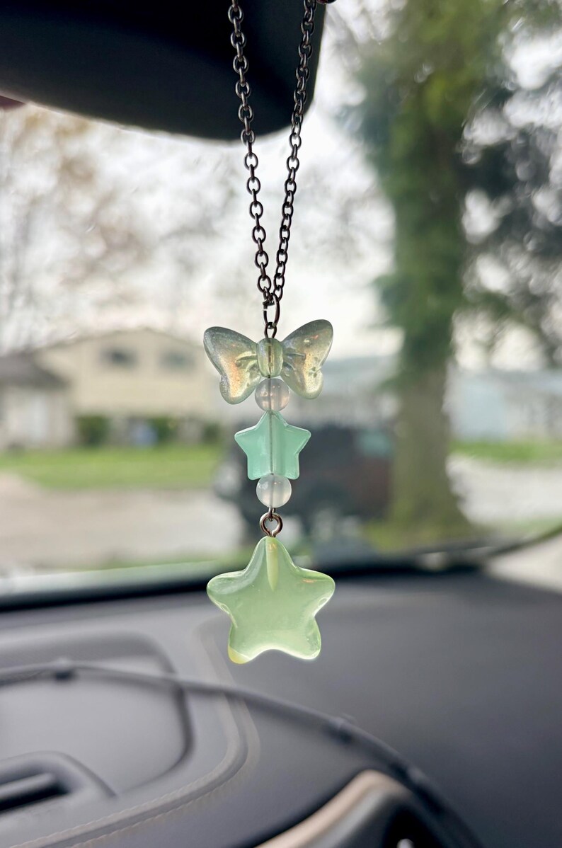 May include: A car charm featuring a chain with a butterfly, star, and bead design. The charm has a light green and clear color scheme. The charm is hanging in front of a car dashboard.