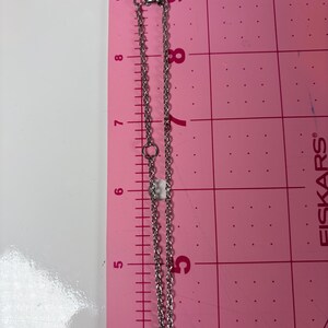 May include: A silver-colored necklace with a pink charm, displayed on a pink surface with a ruler. The necklace chain is approximately 9 inches long. The clasp and chain links are visible.