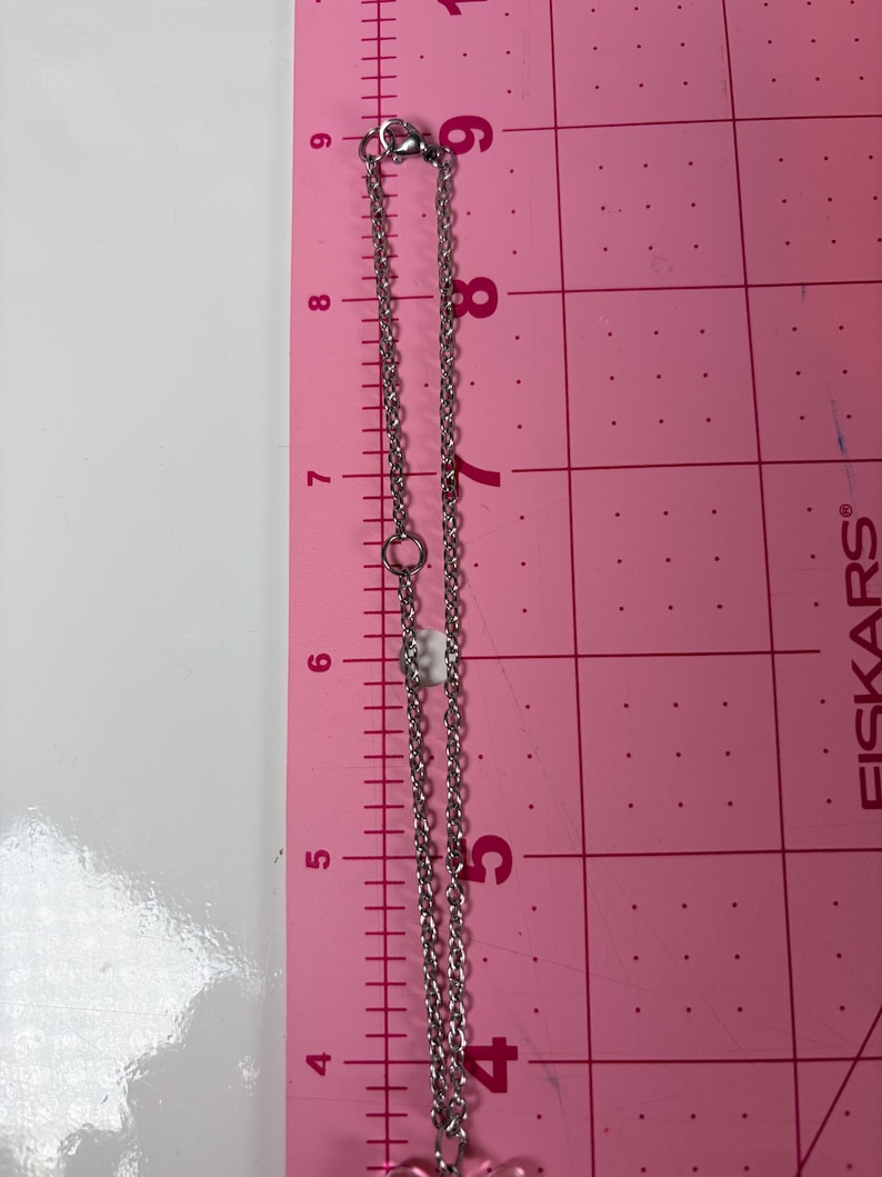 May include: A silver-colored necklace with a pink charm, displayed on a pink surface with a ruler. The necklace chain is approximately 9 inches long. The clasp and chain links are visible.